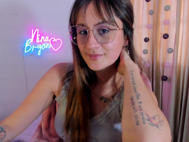 NinaBryons with glasses, smiling, brown hair, wearing a gray tank top, showing a tattoo on her right arm, pink neon sign "Nina Bryans" on the left.