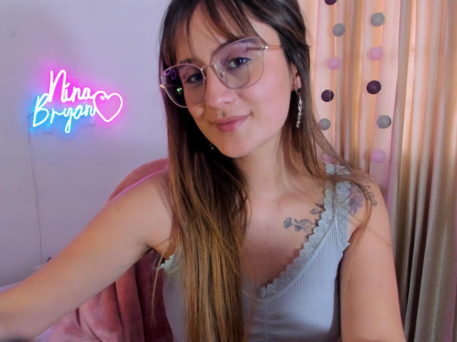 NinaBryons with long brown hair, glasses, and a gray lace-trimmed tank top, showing a tattoo on her right shoulder, smiling at the camera. Boobs are visible.