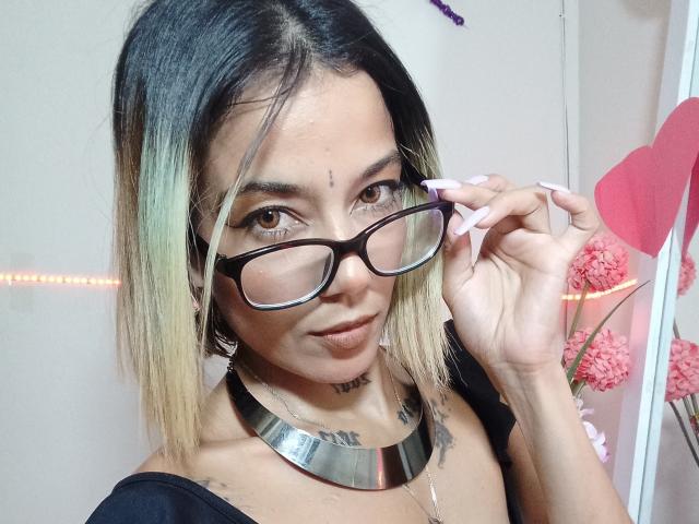 Veroo69 with medium-length blonde and black hair, wearing black glasses, metallic necklace, and black top, showing off long pink nails, has a forehead bindi, and tattoos.