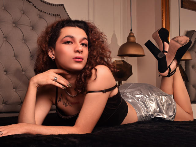 CrystalCraze with curly brown hair, black strap top, and shiny silver shorts, lying on a black bed, wearing black high heels, showing tattoos on her hand and chest.