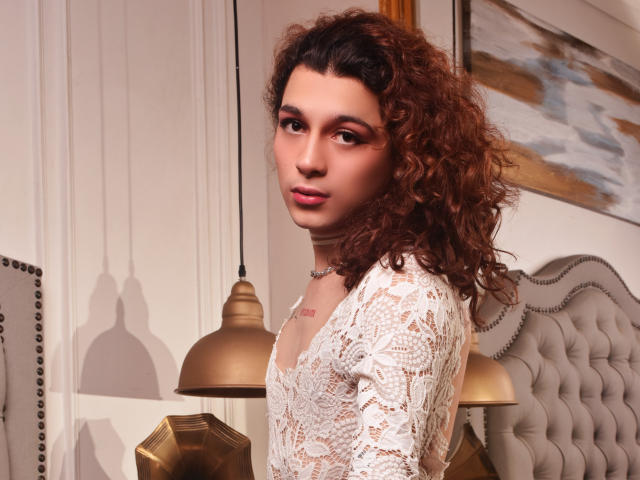 CrystalCraze has curly brown hair, wearing a white lace top, showing a cute face with pink lipstick, and a small tattoo on the chest. She's looking over her shoulder.