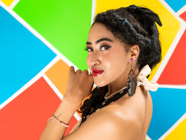 MichellDiaz has dark curly hair, red lipstick, and black tassels in her ears. She's smiling with her hand near her face, showing off her nose ring and bracelet.