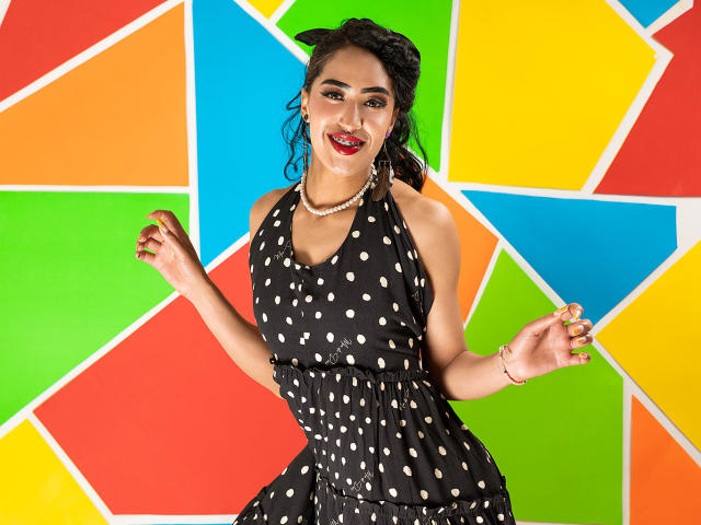 MichellDiaz has dark skin, red lipstick, and black polka dot dress with halter neck, pearl necklace, and curly hair. She's smiling, hands raised, wearing green nail polish.