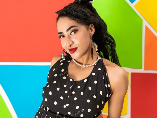 MichellDiaz has dark skin, red lipstick, and black polka dot dress, wearing a pearl necklace and earrings, with curly black hair and a confident smile.