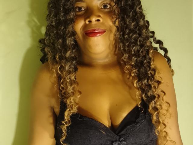 ElysiaX has curly brown hair with blonde tips, wearing a black lace top, showing her large, rounded boobs, and smiling with red lipstick.