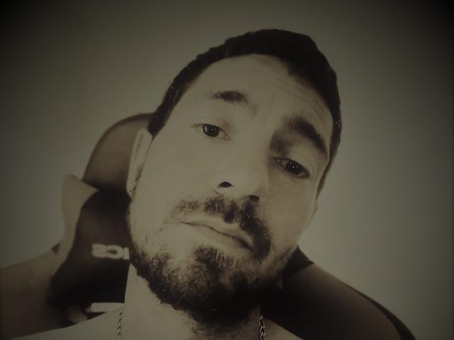 Freya69s has a slightly tired expression, short dark hair, a beard, and a chain necklace. A blurry chair is behind her head. Sepia-toned image.