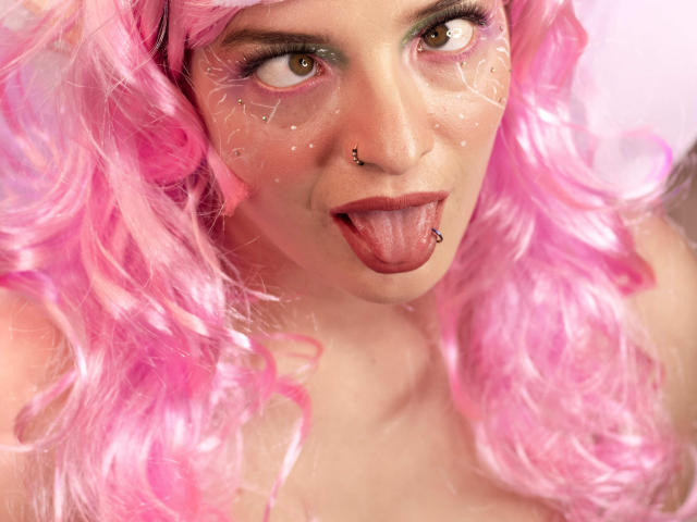 TeddyAndHottie con cabello rizado rosa, ojos marrones y maquillaje en la cara, sacando la lengua, con piercings en la nariz y la lengua, luciendo un aspecto juguetón. Los senos no son visibles. Fondo rosa y blanco suave..