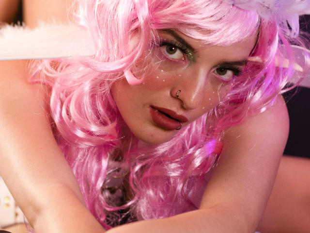 TeddyAndHottie con capelli ricci rosa, piercing al naso e glitter sul viso, guarda verso l'alto con la lingua fuori, ha una piuma bianca tra i capelli e indossa una maglietta rosa..