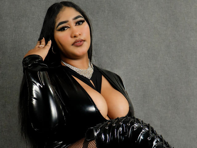 MelisaJackie with big boobs, wearing a black shiny latex top, diamond choker, and black gloves, has long black hair, heavy eyeliner, and a confident expression.