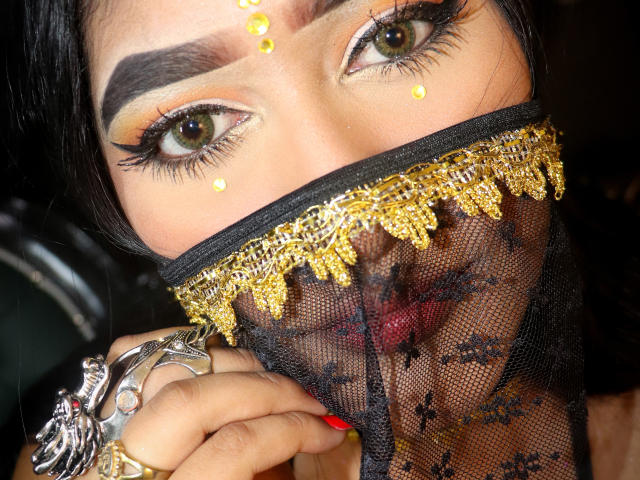 MelisaJackie's face with dark hair, green eyes, and gold eyeliner. She's wearing a black mesh mask with gold trim, showing red lips. She's got silver rings and red nails.