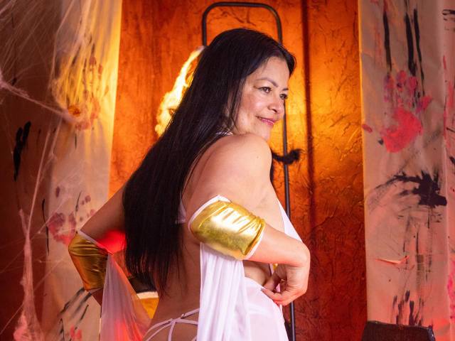 AnaMaturee, com longos cabelos pretos, braceletes dourados, blusa branca transparente e seios visíveis, olhando para trás com um sorriso, em um quarto com iluminação quente e decoração em tons de laranja..