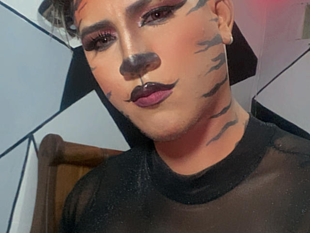 AngelFloress has tiger stripes painted on their face, dark purple lipstick, and wears a sheer black top. They have a nose ring and short, curly brown hair.
