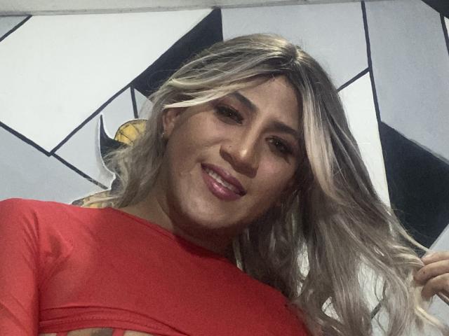 AngelFloress with long blonde hair, red top, smiling at camera, showing her shiny pink lips, holding her hair on the right. Her tits are slightly visible under the top.