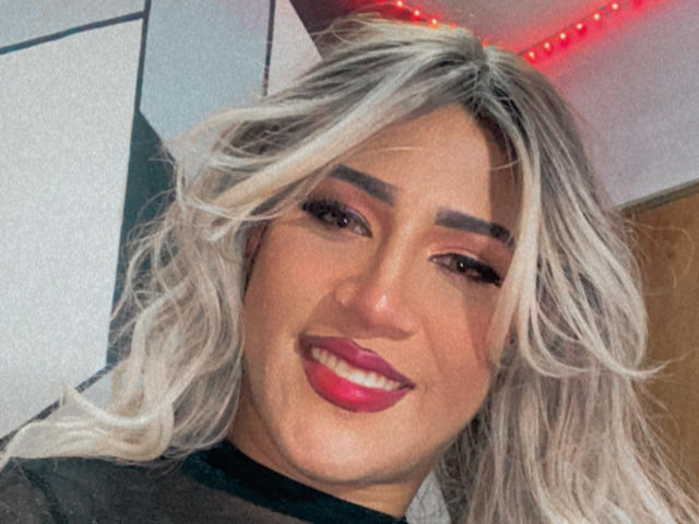 AngelFloress with blonde wavy hair, dark eyebrows, pink lipstick, smiling, wearing a black top, red LED lights in the background.