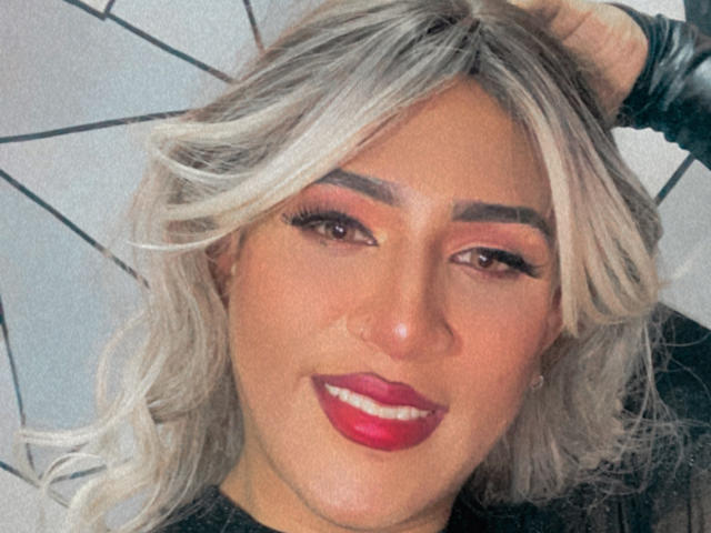 AngelFloress has blonde hair, dark eyebrows, red lipstick, and is smiling with her hand in her hair. She's wearing a black top and has long eyelashes.
