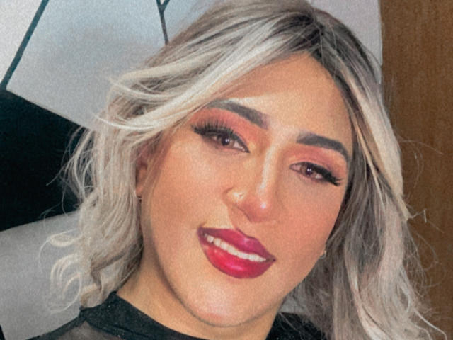 AngelFloress has blonde hair, red lipstick, and pink eye makeup. Her eyebrows are thick, and she's smiling with long eyelashes. She's wearing a black top.