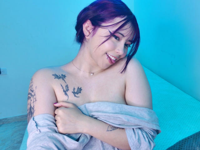Arieyaara with purple hair, smiling, topless, covering her tits with a gray shirt, showing bird tattoos on her chest and arm, in a blue-lit room.