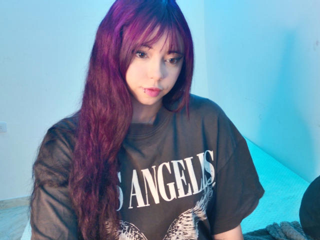 Arieyaara, avec ses longs cheveux violets, son t-shirt noir et son anneau de nez, regarde vers le bas avec un léger sourire, assise dans une pièce éclairée en bleu, et affiche un visage à la fois mignon et séduisant..