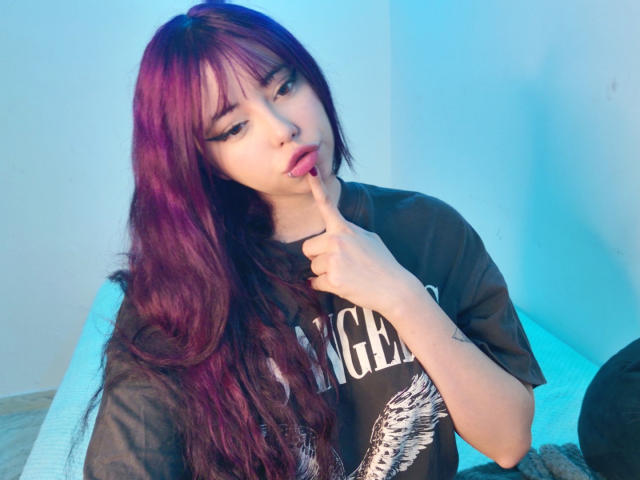 Arieyaara with purple hair, pouty lips, and eyeliner, sucks on her finger, wearing a black T-shirt with a white graphic, in a blue-lit room.