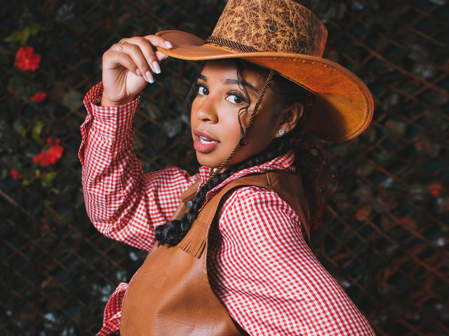 LisaMendez porte un chapeau de cowboy marron, une chemise à carreaux rouges et un gilet beige. Elle regarde par-dessus son épaule avec ses tresses foncées, montrant son joli visage et son petit nez fin..