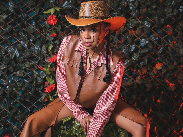 LisaMendez with brown skin, wearing a cowboy hat, checkered shirt with fringe, no pants, sitting with legs spread, showing small tits, against a leafy, red-flowered background.