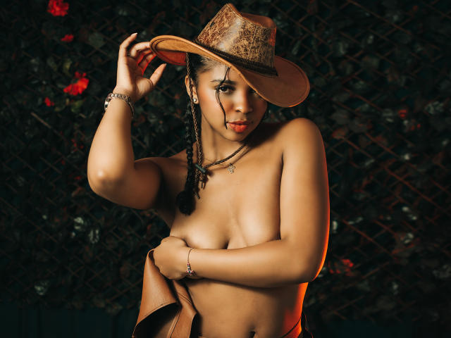 LisaMendez, topless, with brown cowboy hat, holding it, her hand covering her tits, dark braided hair, bracelet, necklace, dark skin, sexy, red lips, dark background, evening light.