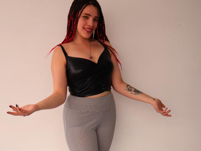 ElizaLovely with red and black braided hair, black leather top, and checkered pants, smiling, arms out, showing tattoo on right arm, standing against plain background.