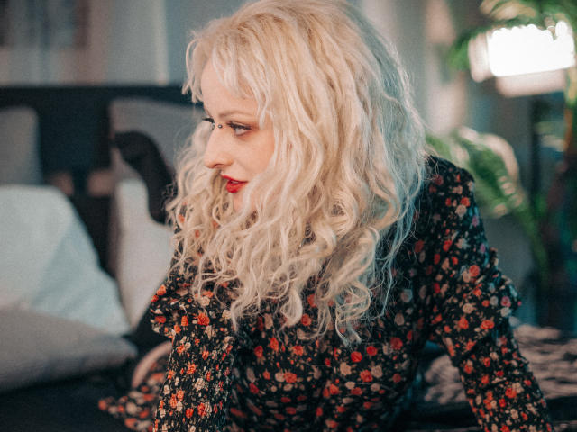NateLove has wavy blonde hair, red lipstick, and a floral dress, looking to the side with a slight smile. Boobs partially visible. Soft, natural light.