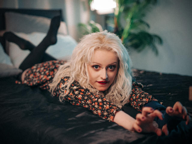 NateLove with blonde curly hair, lying on black bed, wearing floral top, black sleeves, and black stockings, staring at camera, arms outstretched, red lipstick, bedroom setting.