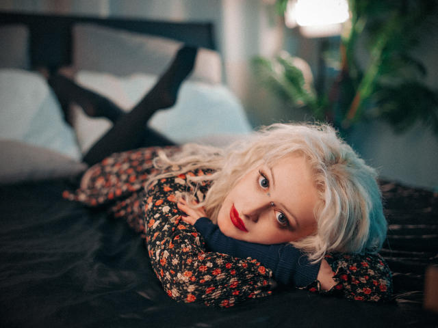NateLove with platinum blonde hair, red lipstick, floral sweater, and black socks, lying on a bed, head on hands, legs crossed, looking at camera with a sexy, relaxed expression.