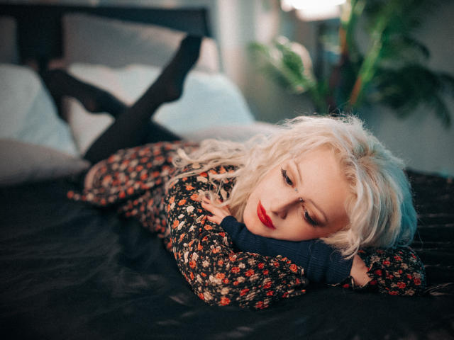 NateLove with platinum blonde hair, lying on a bed, wearing a floral sweater, black tights, and red lipstick, head on dark pillow, legs up, looking down, soft light.