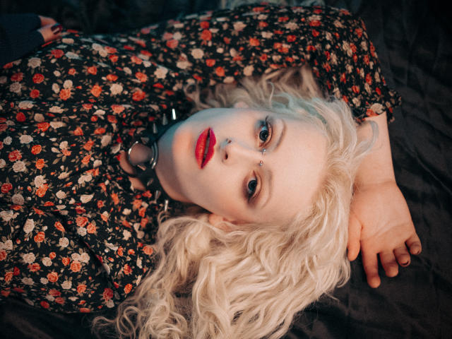 NateLove with pale skin, blonde curly hair, red lipstick, floral shirt, black choker, lying on dark fabric, arms behind head, looking up with blue eyes, slightly parted lips.