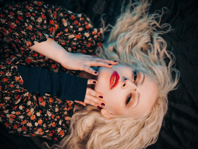 NateLove has wavy blonde hair, red lipstick, and black floral dress, lying down with hands near face, showing dark nail polish, nose ring, and dark blue turtleneck.