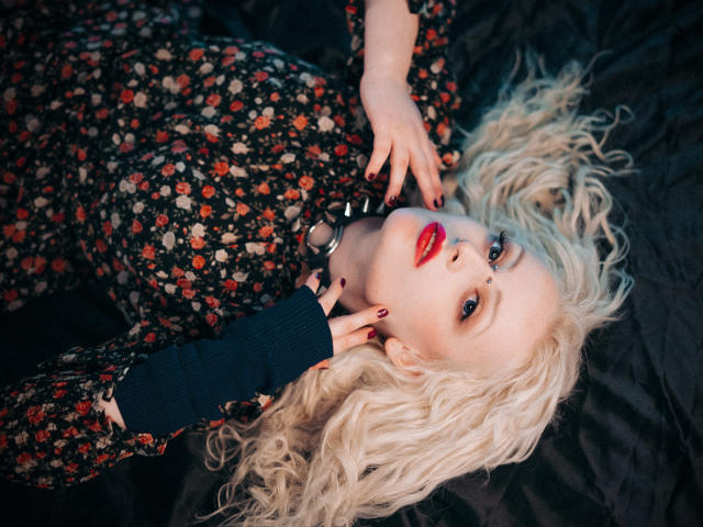 NateLove with curly blonde hair, red lips, floral dress, and dark nails, lying on black fabric, touching her neck, wearing a blue sleeve, looking up with a sexy expression.