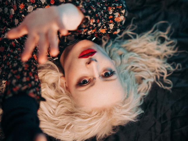 NateLove with curly blonde hair, red lipstick, and floral shirt, reaching out with hand in focus, blue eyes, nose piercing, lying on dark surface, intense gaze.