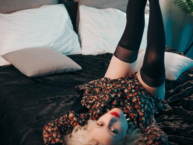 NateLove is lying on a bed with legs up, wearing a floral dress and black thigh-high socks, showing her bare ass. Her red lipstick is prominent.
