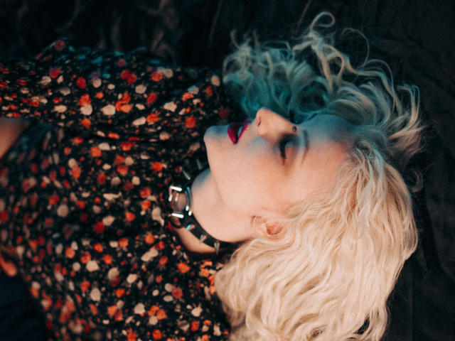 NateLove with curly blonde hair, red lips, and a floral shirt, lying down with eyes closed, wearing a black spiked choker, looking sexy and relaxed.