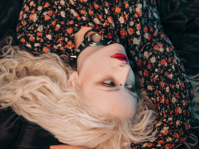 NateLove with pale skin, blonde curly hair, red lipstick, floral dress, black cuffs, lying down, eyes half-open, looking up, nose piercing, dark background.