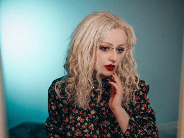 NateLove has wavy blonde hair, red lipstick, and a floral dress, with a hand near her face, looking seductive with a slight smile. Her tits are partially visible.
