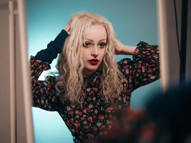 NateLove with wavy blonde hair, red lipstick, floral dress, glasses, hands in hair, blue background, looking serious, wearing black sleeves, reflected in mirror, slightly blurred.