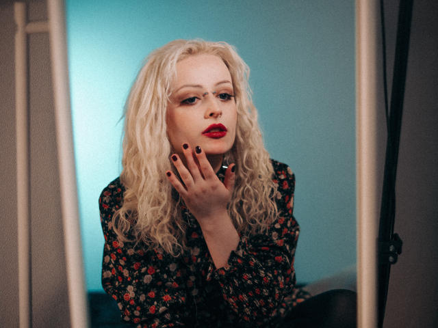 NateLove with curly blonde hair, red lipstick, floral shirt, black nails, hand on cheek, looking into mirror, blue background, soft lighting, slightly seductive expression.