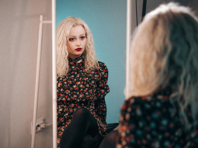 NateLove with curly blonde hair, wearing a floral shirt, black pants, and red lipstick, sits in front of a mirror, looking at herself with a slightly serious expression.