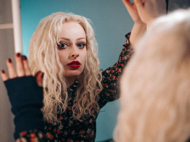 NateLove with curly blonde hair, red lipstick, and floral dress, wearing black fingerless glove, staring into mirror, reflection showing her hand. Her tits are visible under the dress.
