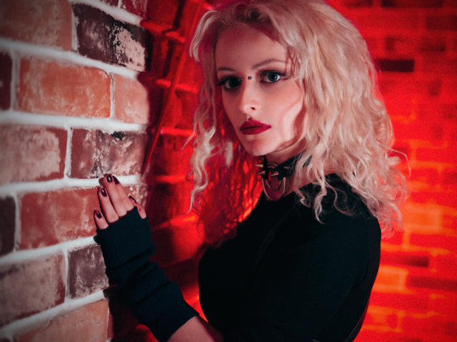 NateLove with blonde curls, black top, fingerless gloves, collar, red lighting, standing against brick wall, looking at camera, hands on wall, red highlights, dark makeup, full lips.