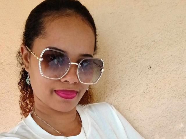 TatianBelle with curly brown hair, wearing white shirt, pink lipstick, and oversized sunglasses with pearl accents, smiling against a textured beige wall.