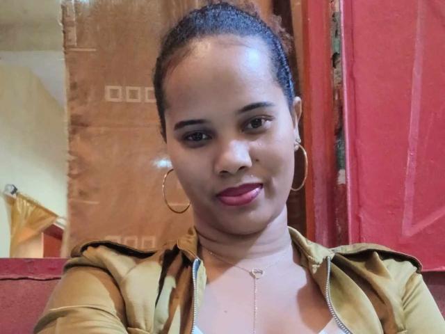 TatianBelle with dark skin, brown eyes, and curly hair in a ponytail, wearing a green jacket, hoop earrings, and pink lipstick, smiling slightly. Her tits are small, visible.