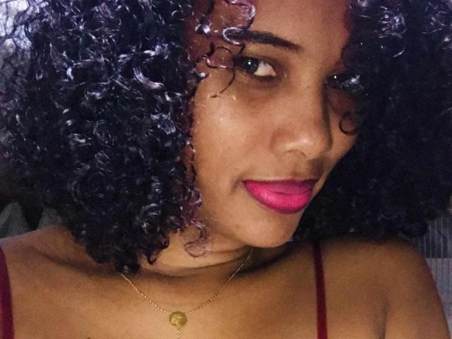 TatianBelle with curly black and purple hair, pink lipstick, and a gold necklace, smiling at the camera, showing her medium brown skin and red bra straps.
