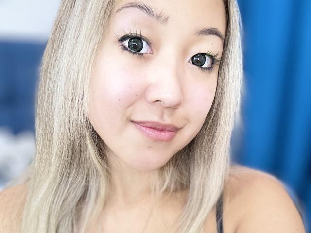 RenyLime has light blonde hair, green eyes, and a small nose. She's smiling with pink lips, wearing black eyeliner, and looking directly at the camera.