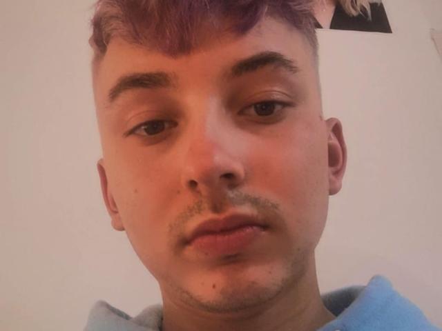 O rosto de Hazuku, com cabelo roxo curto, uma leve barba e uma expressão neutra, vestindo um moletom azul. Suas sobrancelhas são escuras e ligeiramente arqueadas..