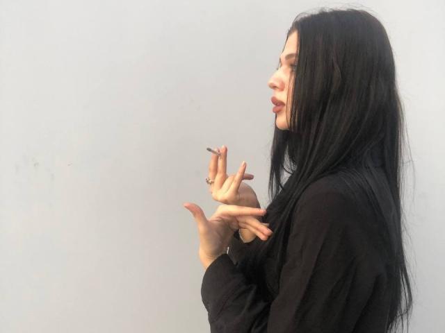 LindaShawna with long black hair, wearing a black top, holding a cigarette in her right hand, ring on her finger, looking side profile, pale skin, relaxed expression.